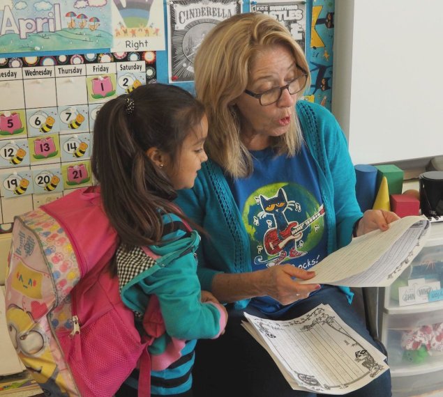 A blonde haired woman with glasses is reading to a little girl with brown hair and a pink backpack
