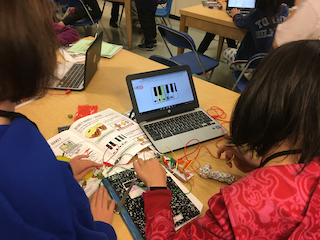 2 students look at a chromebook while touching tin foil and a fork. The foil and fork are attached to wires that are attached to the computer. They are using what is called a Makey Makey to learn what conducts electricity