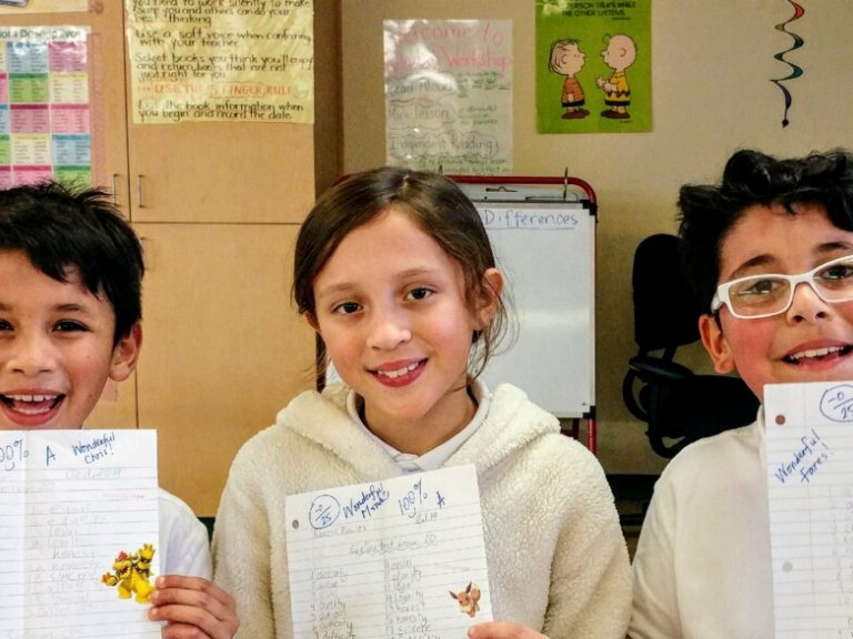 Three elementary students smile in a classroom holding up their test papers.