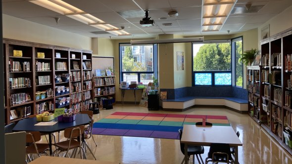 School library with shelves of books, tables for student use, bright windows, and colorful rug.