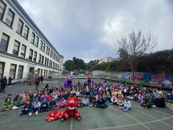 Flynn students K-2, sitting on the blacktop posing with LionDanceMe dragon dancing group