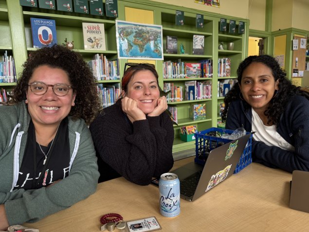 Flynn Kindergarten teachers sitting in library together (Ms. Charlotte, Ms. Sarah & Ms. Anaheli)